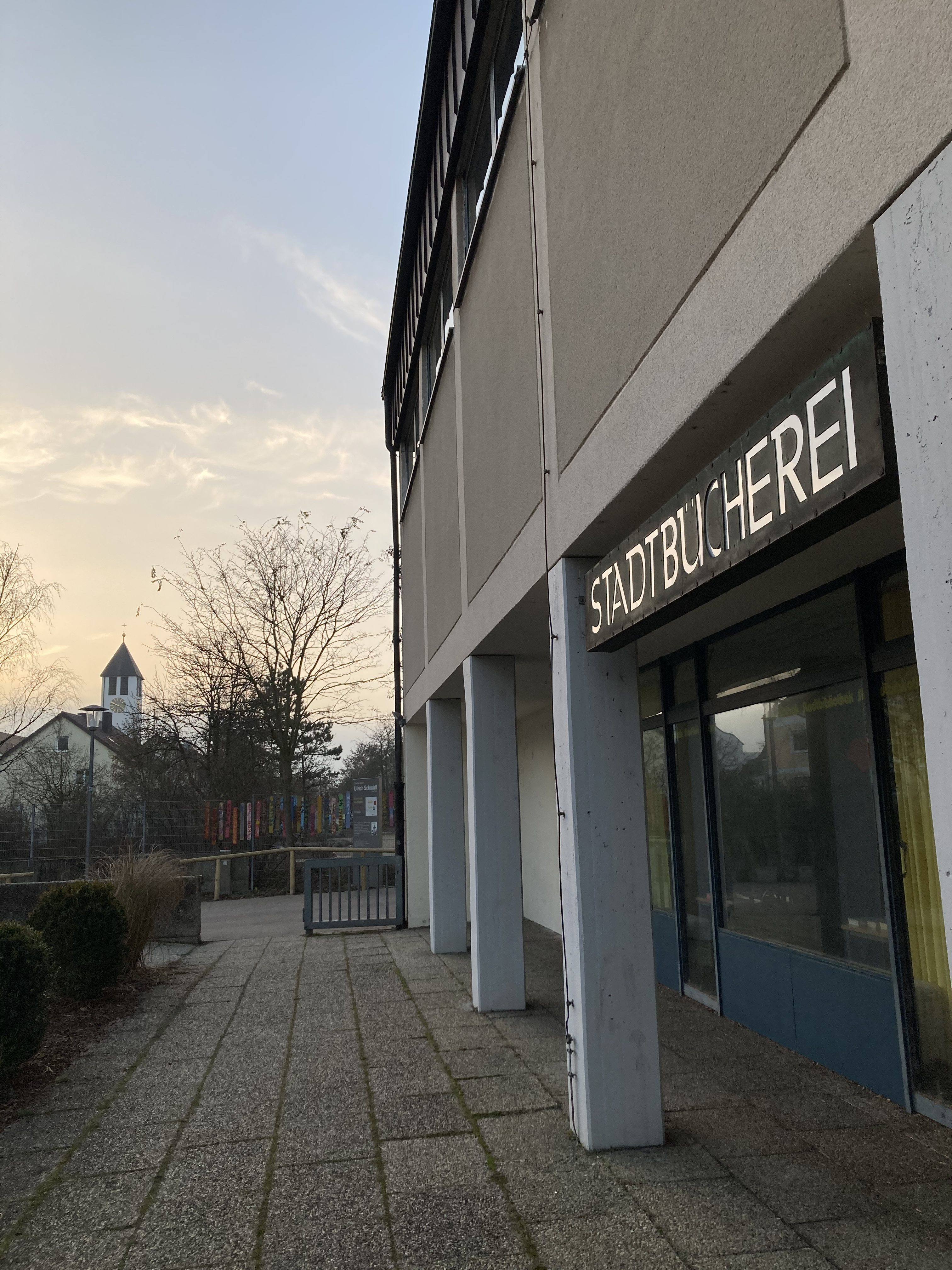 Das Bild zeigt die Außenansicht eines alten Gebäudes mit einem Schild, auf dem "Stadtbücherei" steht, und einem Weg, der an Büschen und einem kleinen Zaun vorbeiführt.