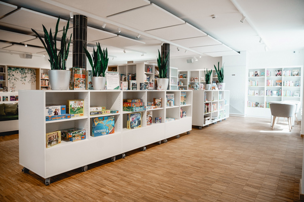Moderne, gut beleuchtete Bibliothek mit weißen Regalen, die mit Spielen und Büchern gefüllt sind, sowie einige große Topfpflanzen und eine gemütliche Sitzecke.
