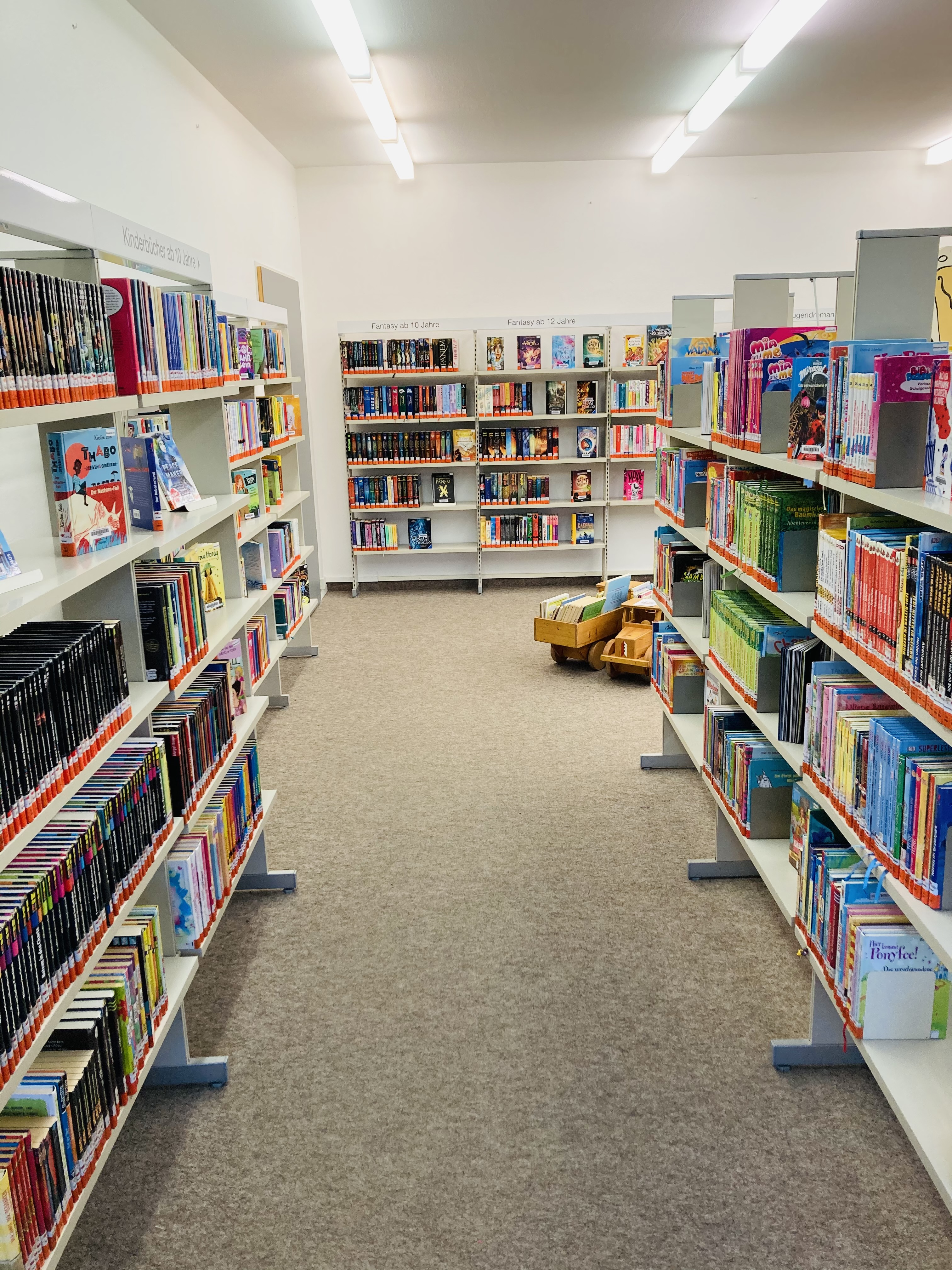 Ein Bücherregalraum in einer Bibliothek mit einem schmalen Gang, in dem auf einem kleinen Spielzeugauto Kinderbücher stehen.
