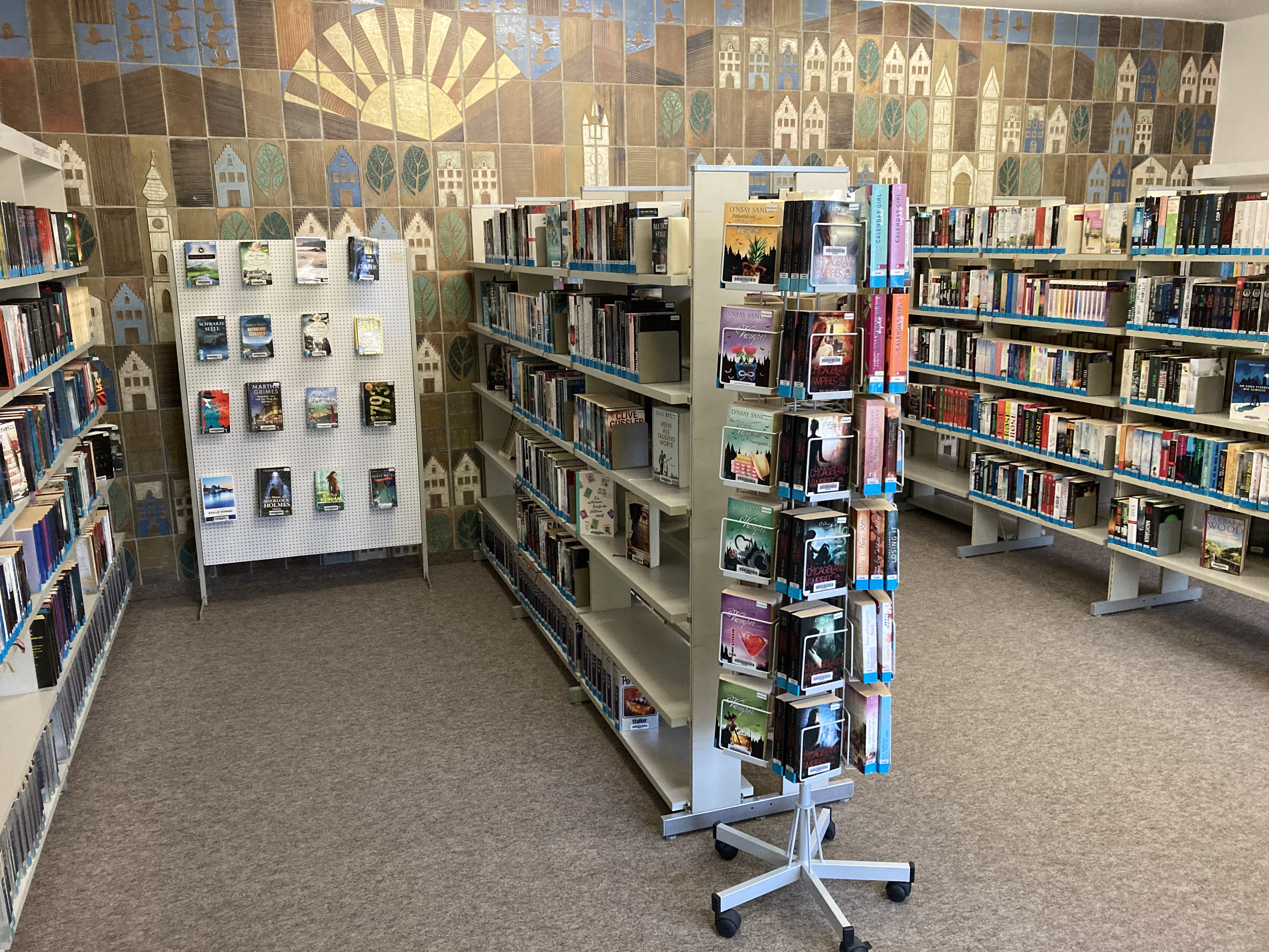 Bücherraum in einer Bibliothek mit mehreren Regalen, einem rotierenden Bücherständer und einer Wand mit dekorativen Fliesen im Hintergrund.