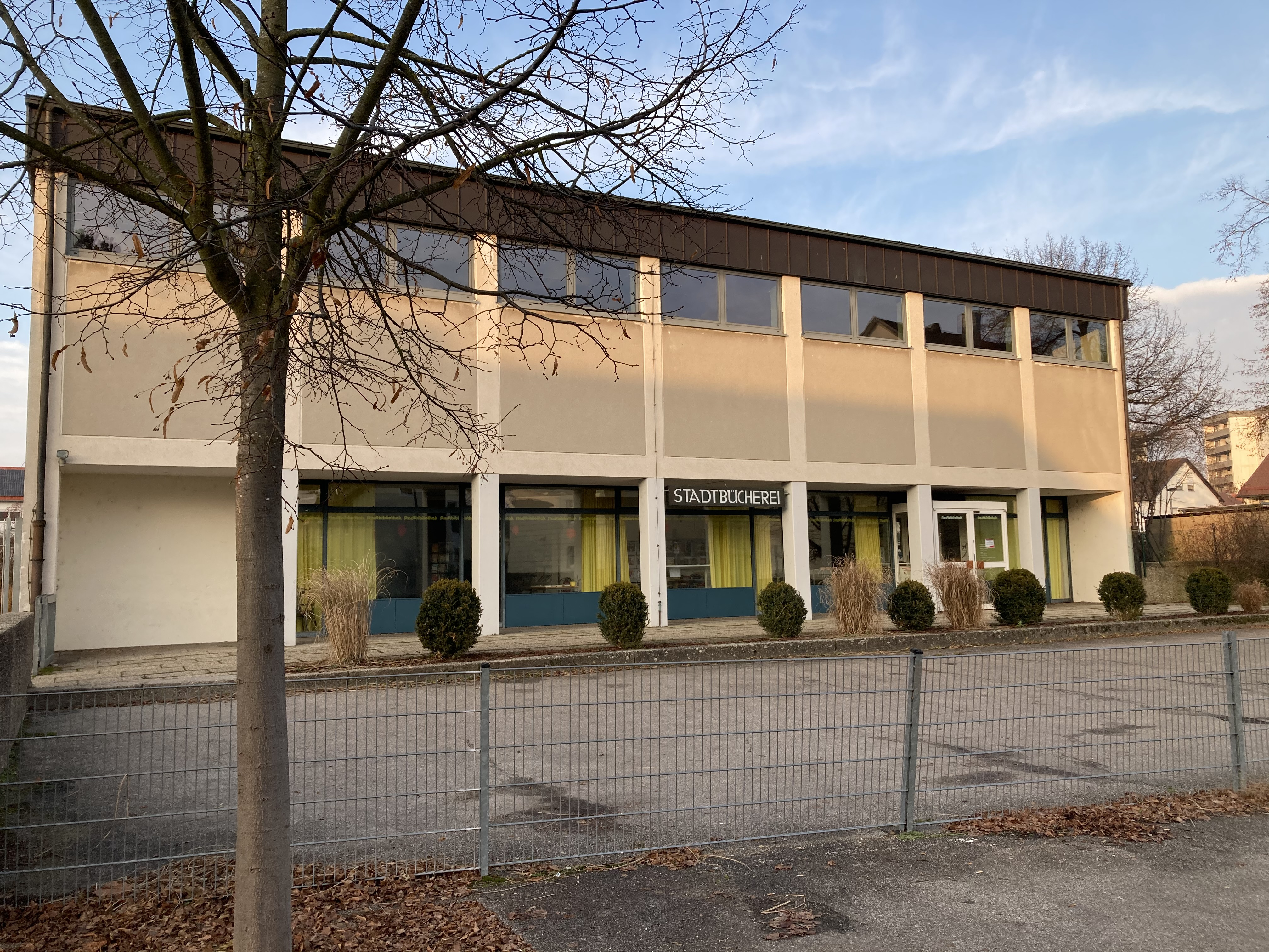 Ein zweistöckiges altes Gebäude mit der Aufschrift "Stadtbücherei", das vor einem kleinen, umzäunten Parkplatz liegt und von Bäumen und Sträuchern umgeben ist.